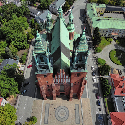 Fotografia przedstawia gotycką ceglaną fasadę frontową katedry poznańskiej. Świątynia posiada dwie wieże, zakończone późnobarokowymi zwieńczeniami i hełmami. Obiekt zlokalizowany jest w otoczeniu drzew i krzewów. Fotografia wykonana została latem - słońce oświetla kościół, niebo jest niebieskie, z niewielką liczbą pierzastych chmur.